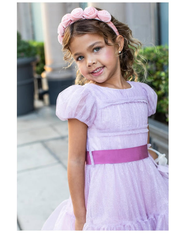 Young girl in a lavender dress with a pink belt and lavender rosette velvet headband, standing outdoors.