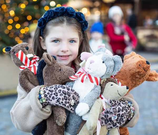 Young girl  wearing navy blue rosette headband holding a collection of stuffed animals with a blurred festive background