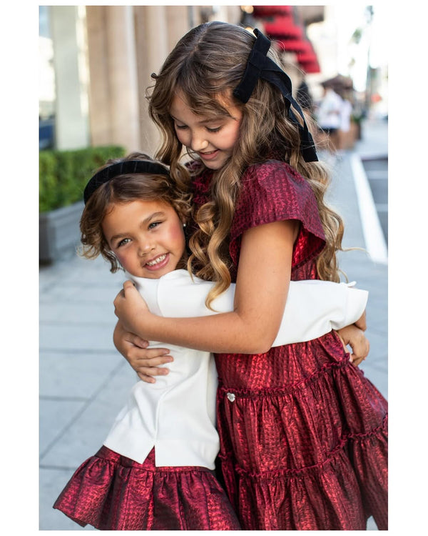 Two young girls in matching red and white dresses hugging on a city street wearing black suede headbands