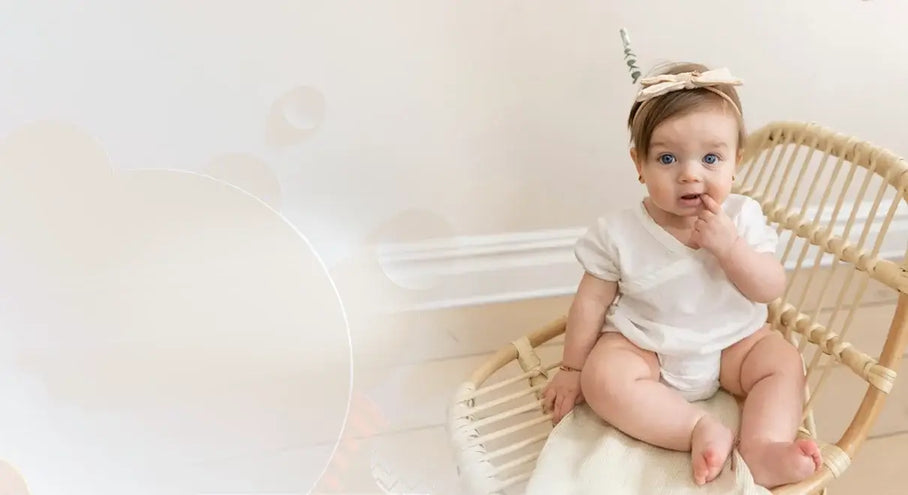 A baby in a white outfit sits on a wicker chair, wearing a headband with a small unicorn horn.