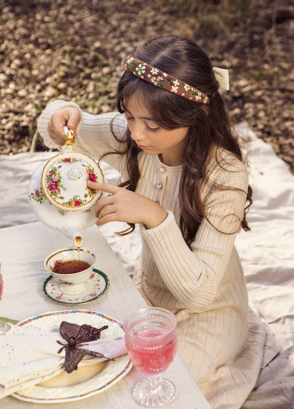 Young girl at a tea party setup outdoors with a teapot and tea cup. Wearing brown embroidered headband.