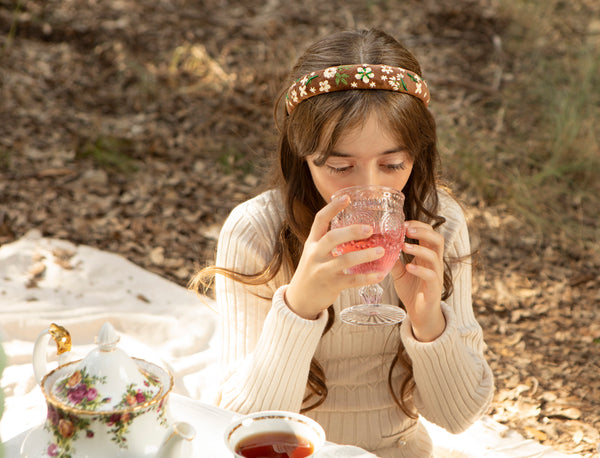 A little girl drinking juice from a glass in a natural setting with a floral hand embroidered headband.